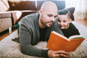 A millennial father and his boy lay on a rug on the living room floor, reading a book together. A good teaching opportunity as the child is learning to read. The dad smiles as he reads, the son looking on attentively.