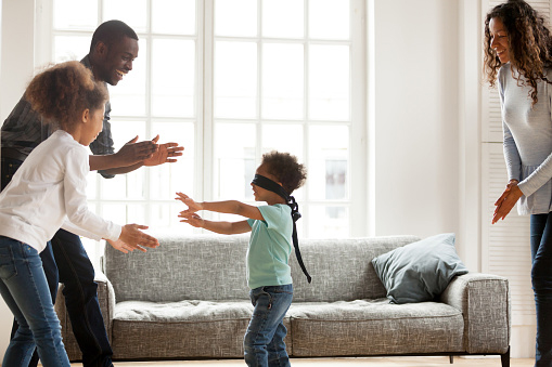 Family Fun Activities: Funny little blindfolded boy play hide and seek game with mixed race family in living room, toddler have fun with black parents and sister, spend time together laughing entertaining at home