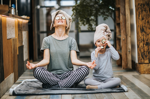 Mother and little girl taking care of their bodies in the morning while doing Yoga meditation exercises at home. Girl is cheating while eating cucumbers from her eyes and looking at camera. self care
