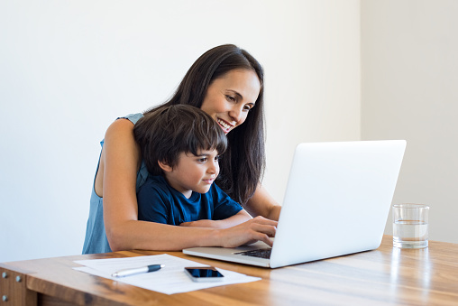 Young mother and son sitting at table and using laptop at home. Smiling mom working at home with her child on the knees while writing an email. Young woman teaching little boy to use the computer.