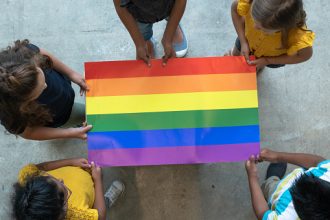 A group of multi-ethnic elementary students stand around a colorful gay pride poster. They are each holding an edge of the poster so it is out flat for the camera to see. Only the tops of their heads can be seen as the picture is taken from an aerial view. Each of the students are wearing casual clothing.