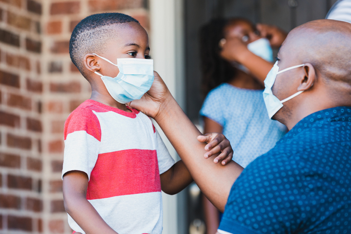 The mid adult father kneels down to adjust his young son's protective mask before going on a walk during the coronavirus epidemic.  In the background, an unrecognizable mom helps her daughter with her mask.