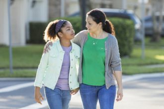 Help Your Child Speak for Themselves: A mature Hispanic woman, in her 40s, walking with her mixed race Hispanic and African-American 11 year old daughter outdoors in their residential neighborhood. Her arm is around her the girl's shoulders as they cross a street. They are looking at each other with serious expressions. Perhaps mom is giving her daughter a pep talk or offering some motherly advice.