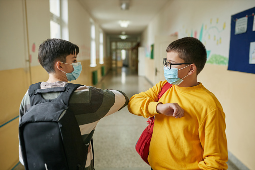 Healthy Habits and Resources: Cheerful elementary students waiting in a school hall before class starts while wearing protective face mask during COVID-19 pandemic