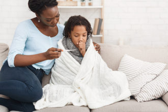 Cold and Flue Season Resources for Parents: Caring African Mom Checking Temperature Of Her Sick Child At Home, Girl In Blanket
