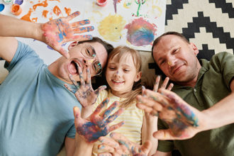 Five DIY Fun Activities with kids - High angle view of happy gay family lying on the floor with their daughter showing their painted hands and smiling they painting at home